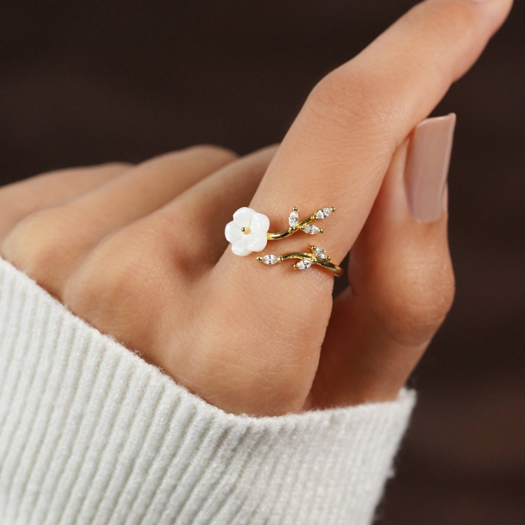 Anel aberto dourado com detalhes de flores e folhas, da marca Rose & Oliver, sobre fundo branco com flores brancas.